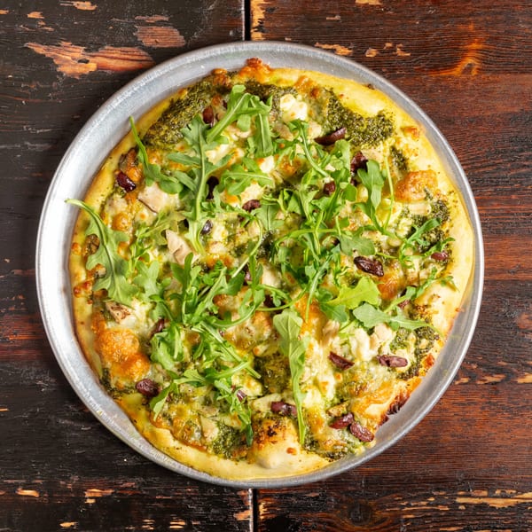 Bambino's pesto chicken pizza with olives, feta, and arugula on a metal pan.