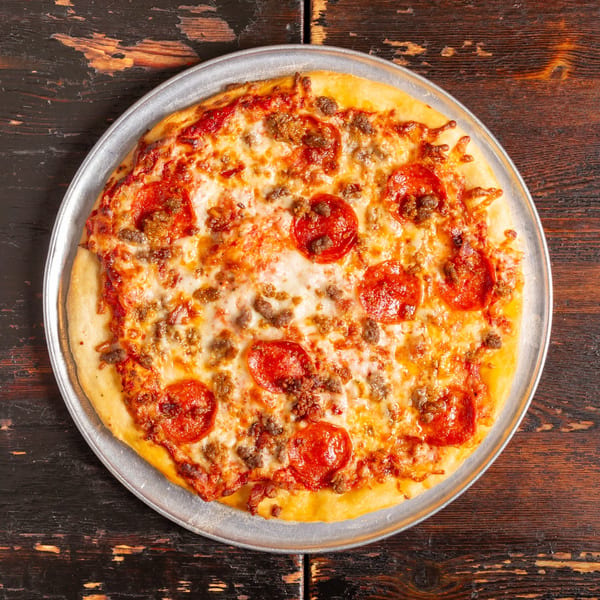 A Bambino's Urban Pizzeria pepperoni and sausage pizza on a metal pan, on a rustic wooden table.