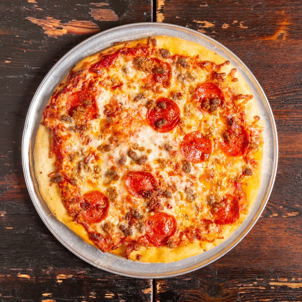 A Bambino's Urban Pizzeria pepperoni and sausage pizza on a metal pan, on a rustic wooden table.