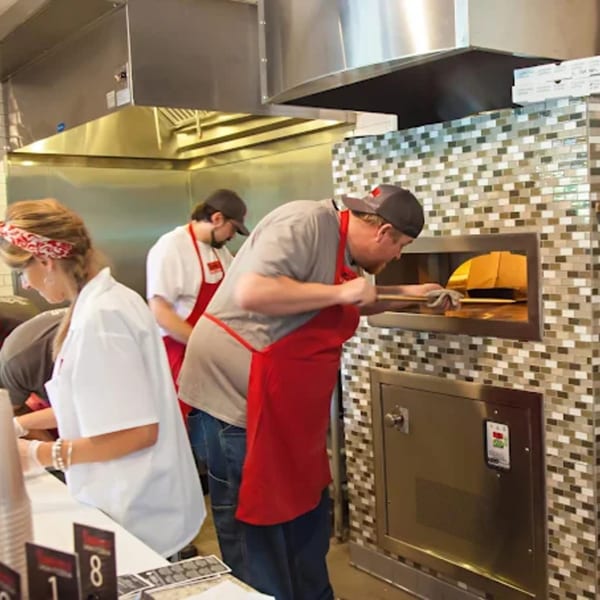Bambino's Urban Pizzeria chefs in red aprons cooking pizzas in a large brick oven.
