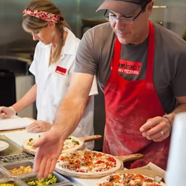Two Bambino's Urban Pizzeria chefs preparing pizzas with various toppings at a counter.